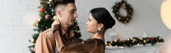 Pareja interracial joven en trajes festivos abrazándose cerca del árbol de Navidad, pancarta - foto de stock