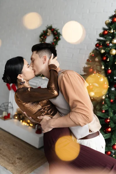 Межрасовая пара в праздничных нарядах целуется, стоя возле елки — стоковое фото