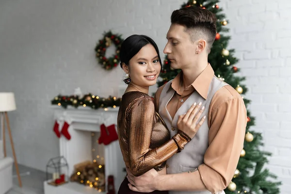 Man hugging cheerful asian wife in festive outfit while standing near Christmas tree — Stock Photo