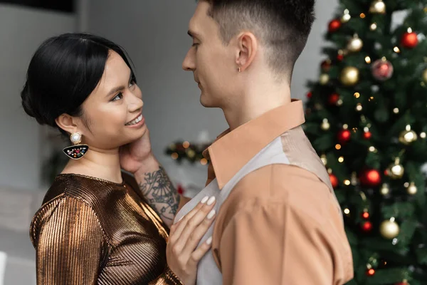 Hombre y alegre asiático mujer en festivo trajes mirando unos a otros cerca de árbol de Navidad - foto de stock