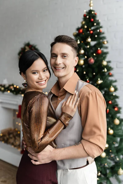 Веселая азиатская женщина и мужчина в праздничных нарядах обнимаются возле елки — стоковое фото