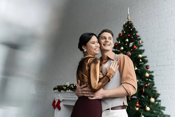 Feliz asiático mujer y hombre en festivo trajes abrazo cerca de árbol de Navidad - foto de stock