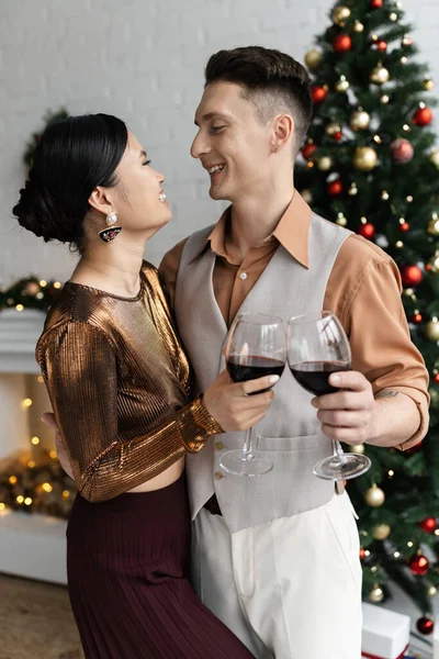 Cheerful multiethnic couple looking at each other while clinking glasses of red wine near Christmas tree — Stock Photo