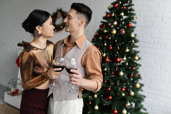 Happy multiethnic couple looking at each other while holding glasses of red wine near Christmas tree — Stock Photo