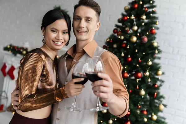 Glückliches multiethnisches Paar mit einem Glas Rotwein in der Nähe des Weihnachtsbaums — Stockfoto