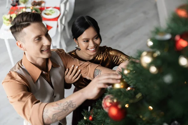 High angle view of smiling interracial couple decorating Christmas tree with shiny baubles — Stock Photo