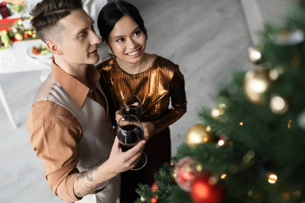 Высокий угол обзора счастливой межрасовой пары в праздничных нарядах, держащей бокалы вина возле елки — стоковое фото