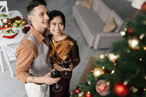Vista de ángulo alto de alegre pareja interracial en trajes festivos sosteniendo copas de vino cerca del árbol de Navidad - foto de stock