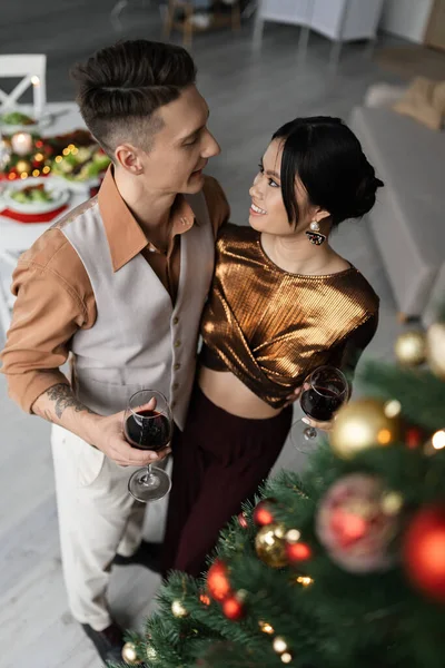 Vista de ángulo alto de feliz pareja interracial en trajes festivos sosteniendo copas de vino cerca del árbol de Navidad decorado - foto de stock