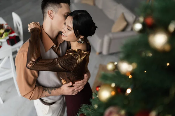 Alto ângulo vista de feliz asiático mulher abraçando tatuado marido perto decorado Natal árvore — Fotografia de Stock