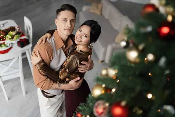 Blick aus der Vogelperspektive auf eine fröhliche asiatische Frau, die sich mit ihrem Mann in der Nähe des Weihnachtsbaums umarmt — Stockfoto