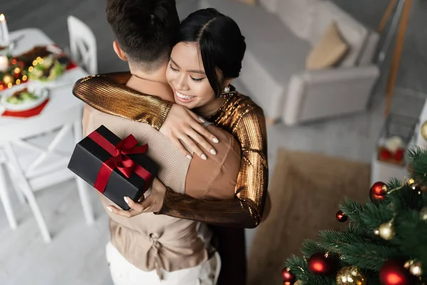 Blick aus der Vogelperspektive auf eine fröhliche asiatische Frau, die ihren Mann umarmt, während sie ein Weihnachtsgeschenk hält — Stockfoto
