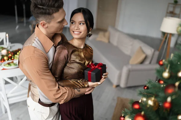 Happy and tattooed man hugging asian wife while holding Christmas present — Stock Photo