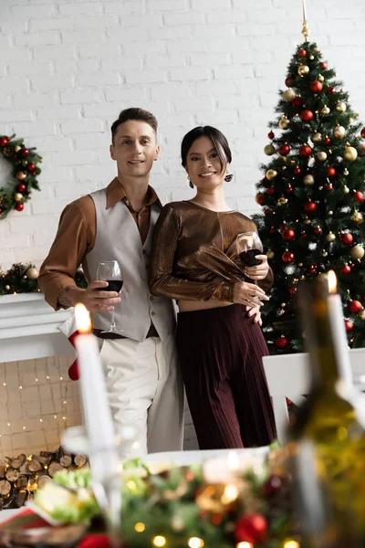 Alegre pareja multiétnica sosteniendo copas de vino durante la celebración de Navidad - foto de stock