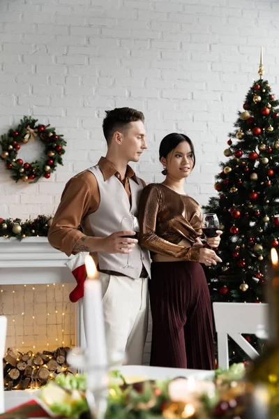 Marido y mujer multiétnicos sosteniendo copas de vino durante la celebración de Navidad - foto de stock