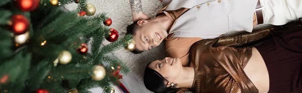 Vista superior de pareja interracial feliz acostado bajo el árbol de Navidad, bandera - foto de stock