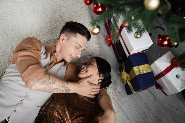 Vista superior de alegre pareja interracial tumbada bajo el árbol de Navidad con regalos - foto de stock