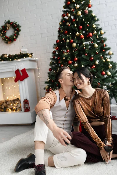 Веселый мужчина целует щеку счастливой азиатской жены, сидя под елкой — стоковое фото