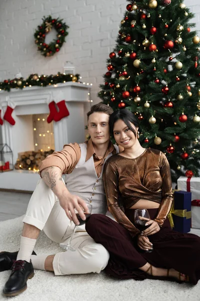 Cheerful man and happy asian woman holding glasses of wine while sitting under Christmas tree — Stock Photo