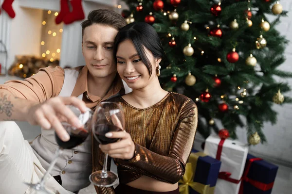 Cheerful man and happy asian woman clinking glasses of wine while sitting under Christmas tree — Stock Photo