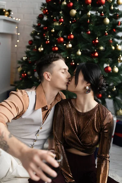 Hombre y asiático mujer con cerrado ojos besándose mientras sentado bajo árbol de Navidad - foto de stock