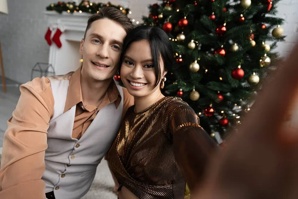 Couple interracial souriant et regardant la caméra tout en étant assis sous l'arbre de Noël — Photo de stock