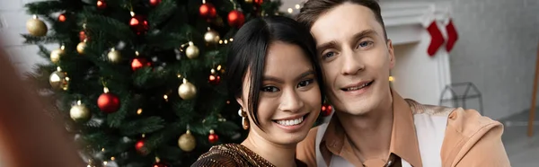Couple interracial souriant et regardant la caméra tout en étant assis sous l'arbre de Noël, bannière — Photo de stock
