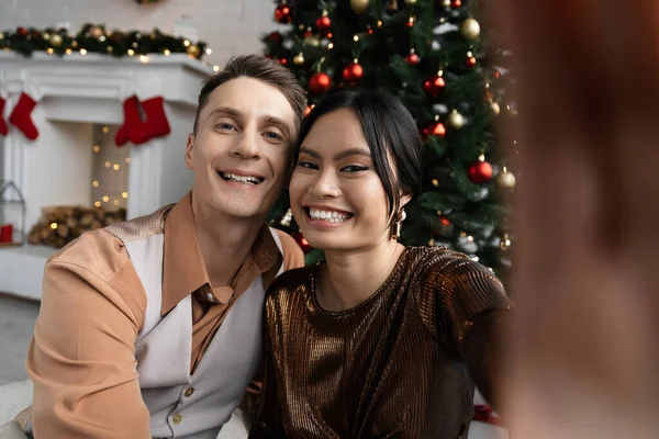 Feliz pareja interracial sonriendo y mirando a la cámara mientras se sienta bajo el árbol de Navidad - foto de stock