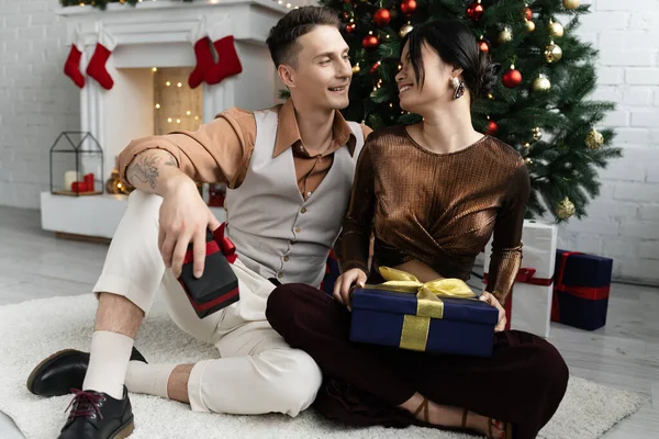 Glückliches gemischtrassiges Paar lächelt und hält Weihnachtsgeschenke in der Hand, während es unter geschmückten Kiefern sitzt — Stockfoto