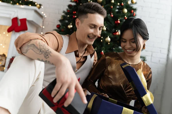 Happy interracial couple opening Christmas present while sitting under decorated fir — Stock Photo