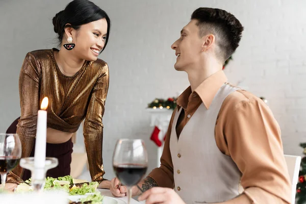 Cheerful interracial couple looking at each other near festive meal and glasses of wine during Christmas — Stock Photo