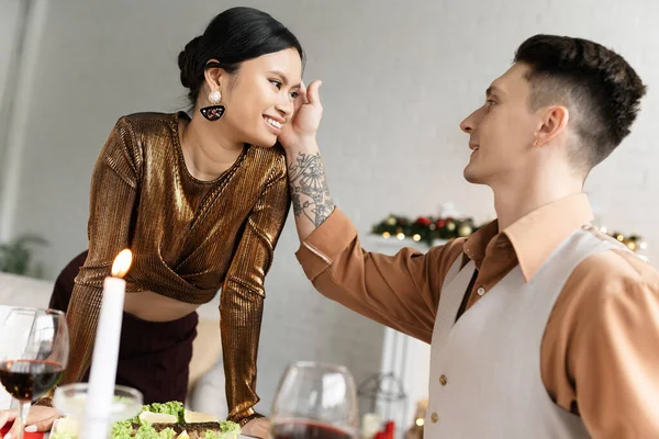 Татуированный мужчина гладит волосы азиатской жены возле праздничного ужина на обеденном столе во время празднования Рождества — стоковое фото
