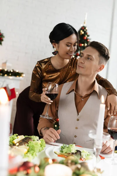 Felice donna asiatica che tiene un bicchiere di vino e abbraccia il marito vicino alla cena festiva durante la celebrazione di Natale — Foto stock