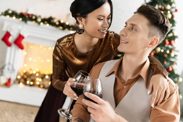 Feliz asiático mujer y alegre hombre tintineo vasos de vino durante la celebración de Navidad - foto de stock