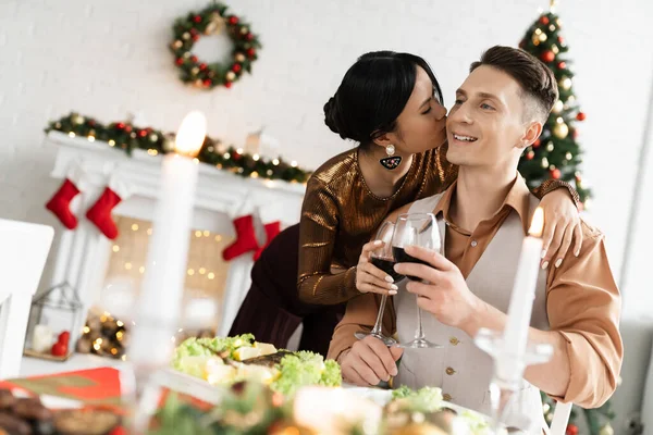 Happy asian woman holding glass of wine and kissing husband near festive dinner during Christmas celebration — Stock Photo