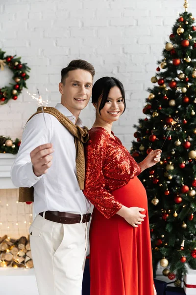 Alegre hombre celebración sparklers con asiático embarazada esposa cerca de navidad árbol - foto de stock