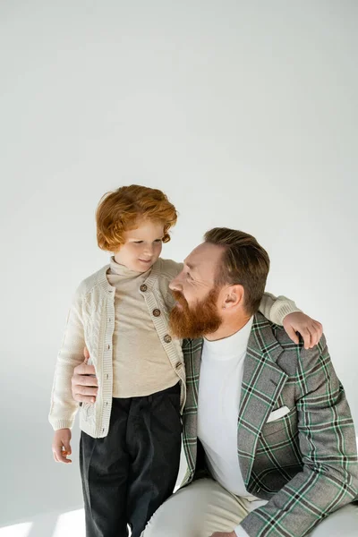 Hombre positivo en chaqueta a cuadros abrazando hijo pelirroja sobre fondo gris - foto de stock