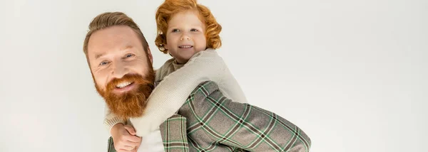 Cheerful redhead boy piggybacking on bearded dad in jacket isolated on grey, banner — Stock Photo