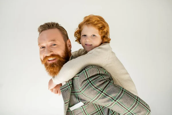 Lächelnder rothaariger Junge huckepack auf Vater in Jacke und Blick in die Kamera isoliert auf grau — Stockfoto
