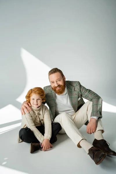 Stylish man hugging redhead son while sitting on grey background with sunlight — Stock Photo