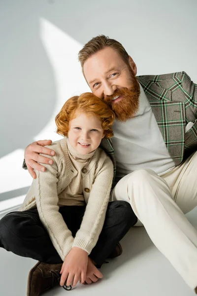 Alegre barbudo homem abraçando vermelho cabelos filho no cinza fundo com luz — Fotografia de Stock