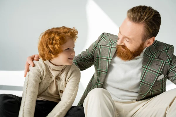 Uomo barbuto sorridente che abbraccia il figlio dai capelli rossi in maglione su sfondo grigio con luce — Foto stock