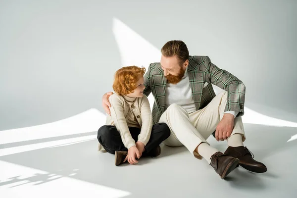 Uomo positivo in giacca abbracciare figlio dai capelli rossi mentre seduto su sfondo grigio con luce — Foto stock