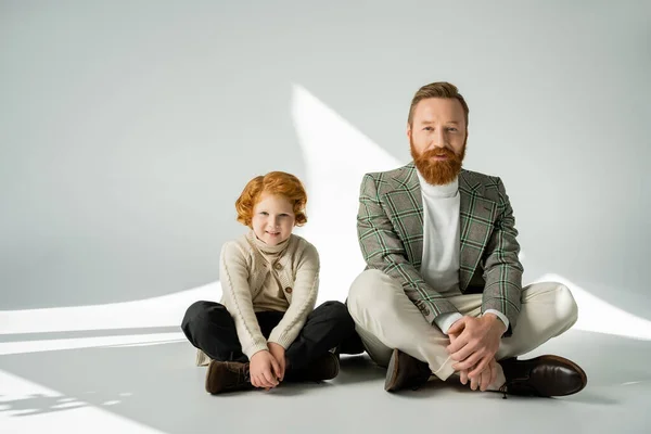 Рудий хлопчик і стильний тато сидить на сірому фоні з сонячним світлом — стокове фото