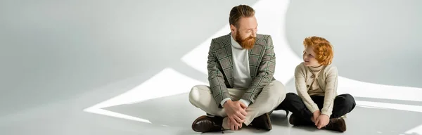 Bearded man in checkered jacket looking at redhead son on grey background with light, banner — Stock Photo