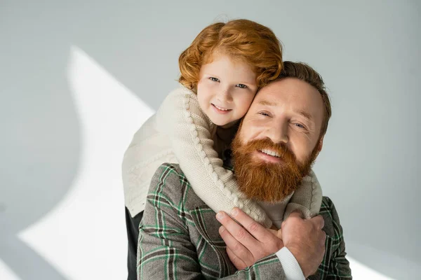 Smiling red haired boy in jumper embracing stylish father on grey background with sunlight — Stock Photo