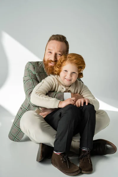 Smiling man in stylish jacket embracing red haired kid on grey background with sunlight — Stock Photo