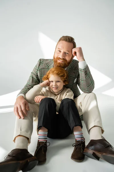 Stylish father and redhead son posing and looking at camera on grey background with sunlight — Stock Photo