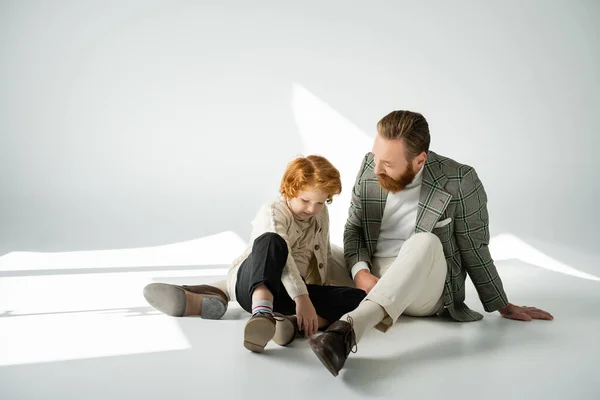 Positive red haired boy sitting near stylish dad on grey background with sunlight — Stock Photo
