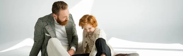 Stylish bearded man in jacket looking at red haired son on grey background with sunlight, banner — Stock Photo